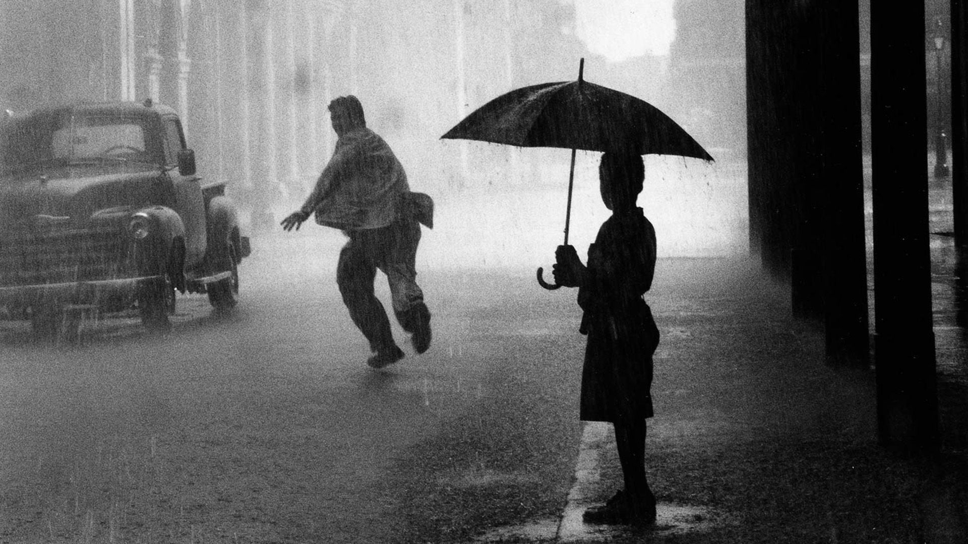 En primer plano, la silueta de un niño bajo un paraguas, observando a un hombre corriendo bajo la lluvia a lo largo de la pared