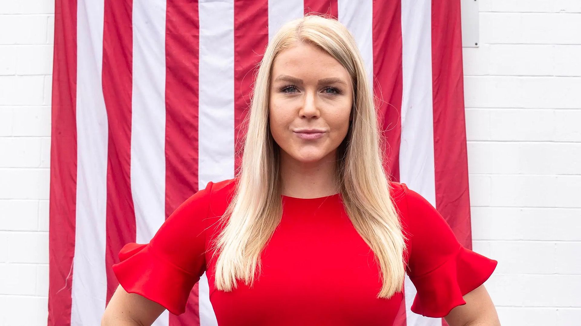 Una rubia de cabello largo con un vestido rojo se encuentra frente al telón de fondo de la bandera estadounidense