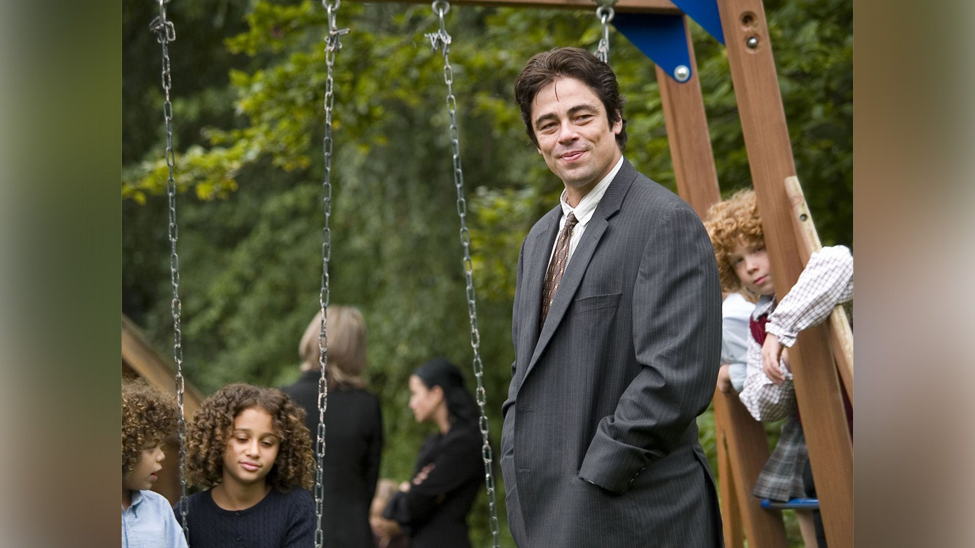 Un hombre sonriente con una chaqueta gris en un parque infantil con varios niños al fondo