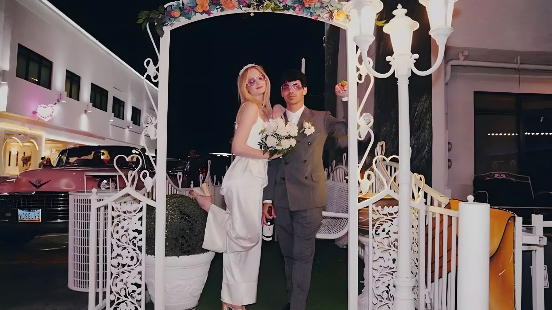 Sophie Turner en un traje pantalón blanco y Joe Jonas en un esmoquin gris bajo un arco de boda