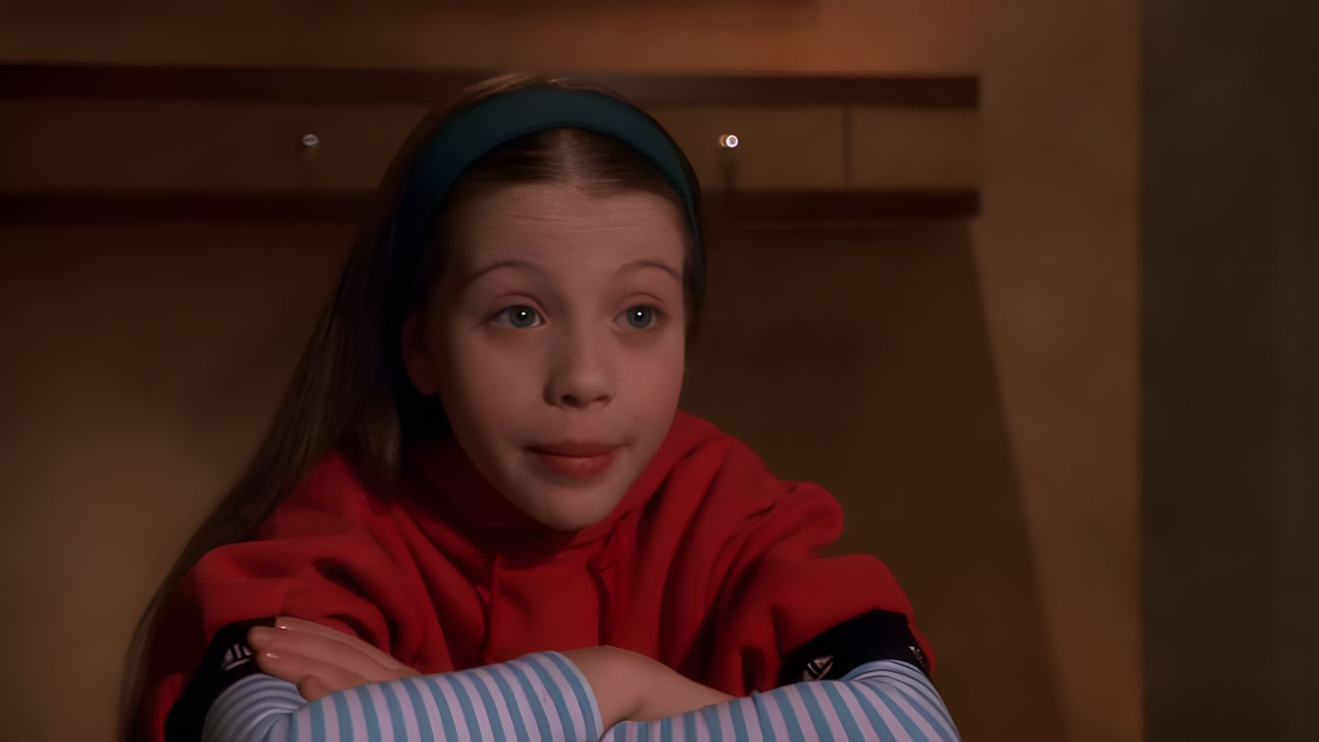 La actriz Michelle Trachtenberg de niña, con un suéter rojo, calentadores de brazos a rayas y una diadema verde