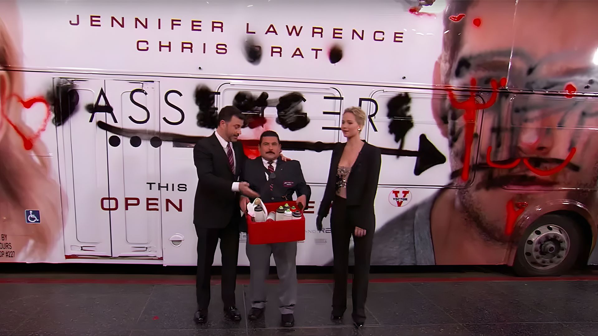 Jimmy Kimmel con un asistente y Jennifer Lawrence están junto a un autobús cubierto de graffiti