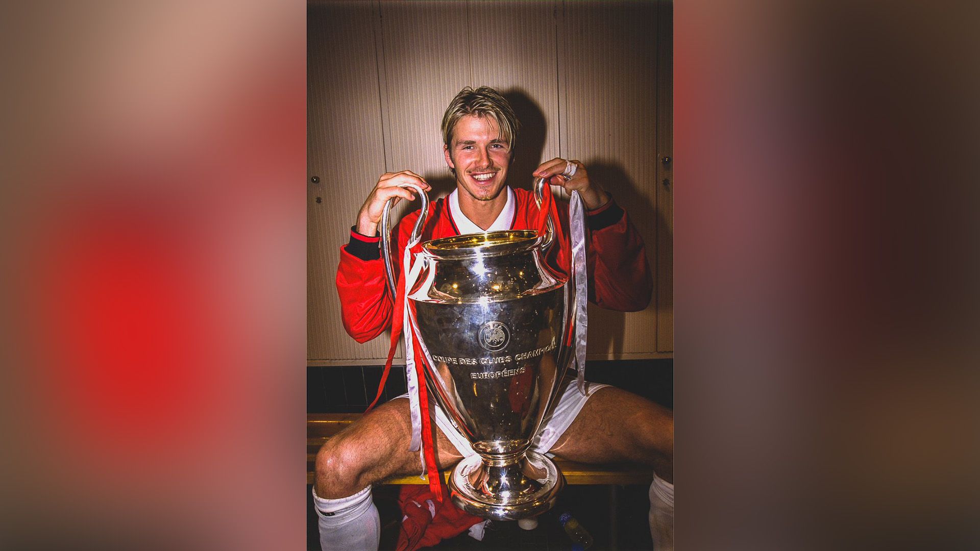 El joven David Beckham sentado en el banco del vestuario deportivo con la Copa UEFA