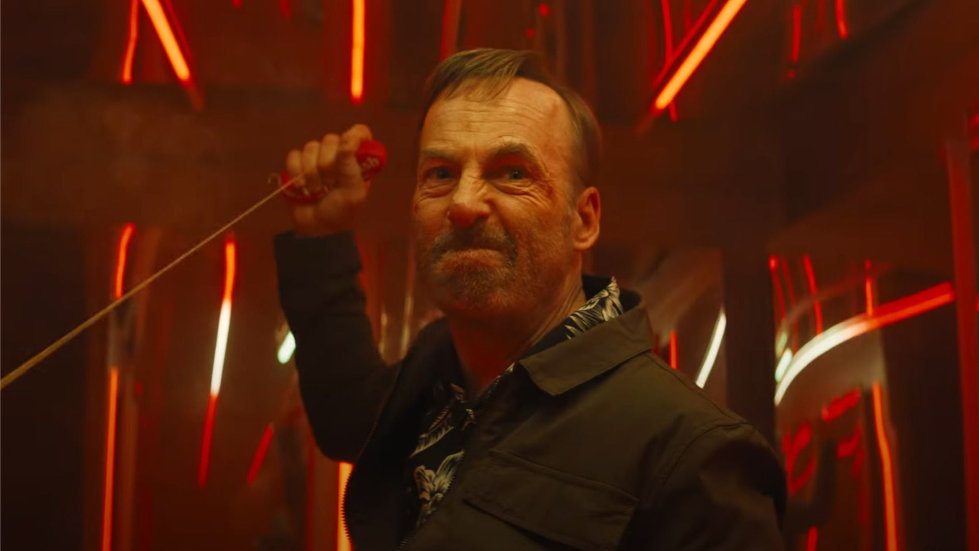 El actor Bob Odenkirk en una chaqueta negra sostiene una cuerda en sus manos y mira enojado hacia la luz roja