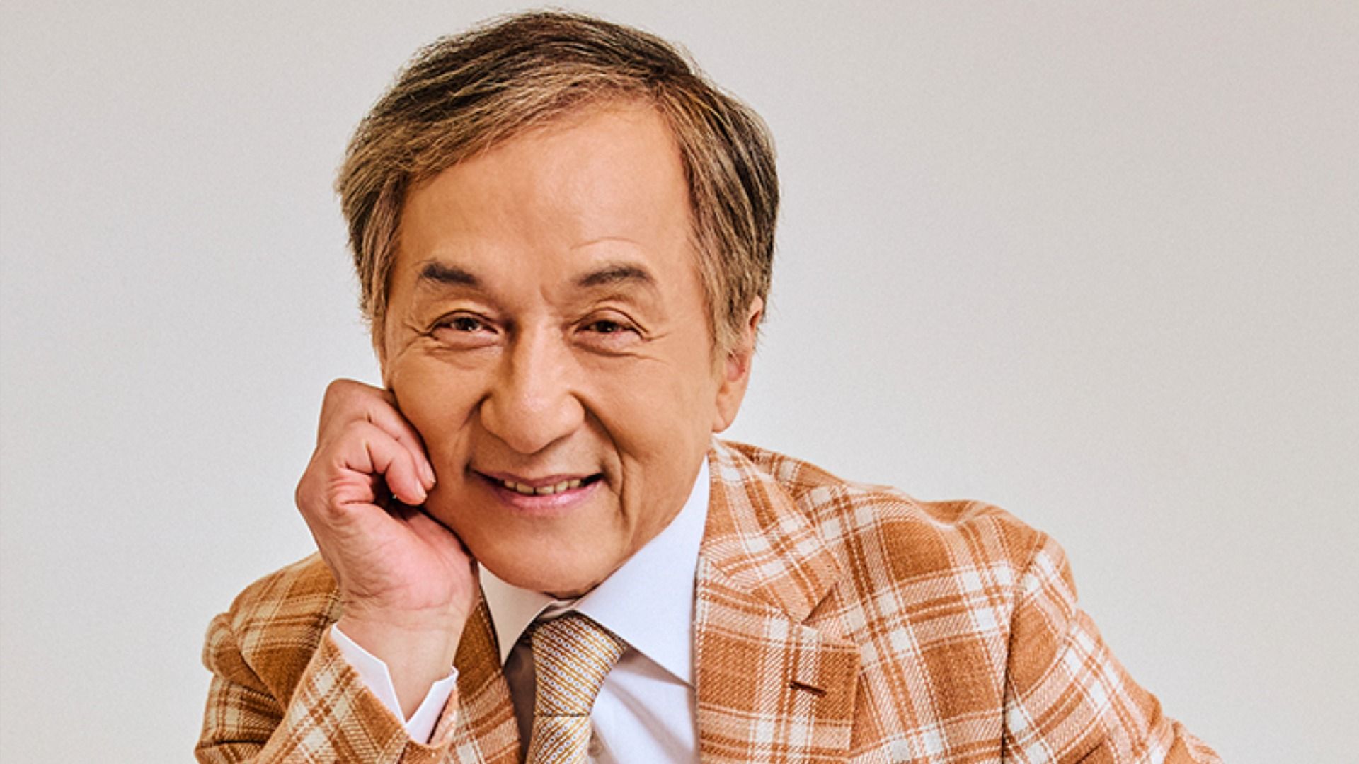 El actor Jackie Chan en una chaqueta marrón a cuadros se sienta, apoyado en su mano, y sonríe a la cámara contra un fondo blanco