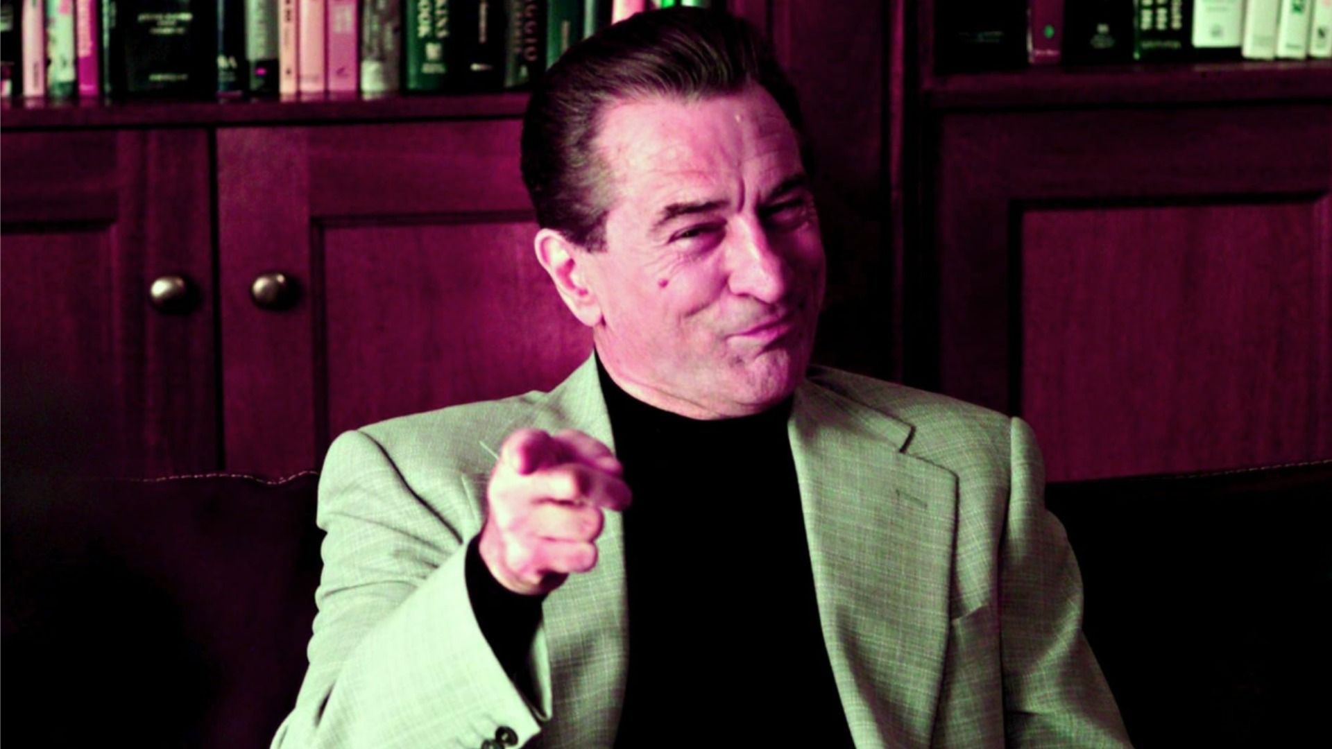 Actor Robert De Niro en una chaqueta gris señala con su dedo índice y sonríe frente a una estantería