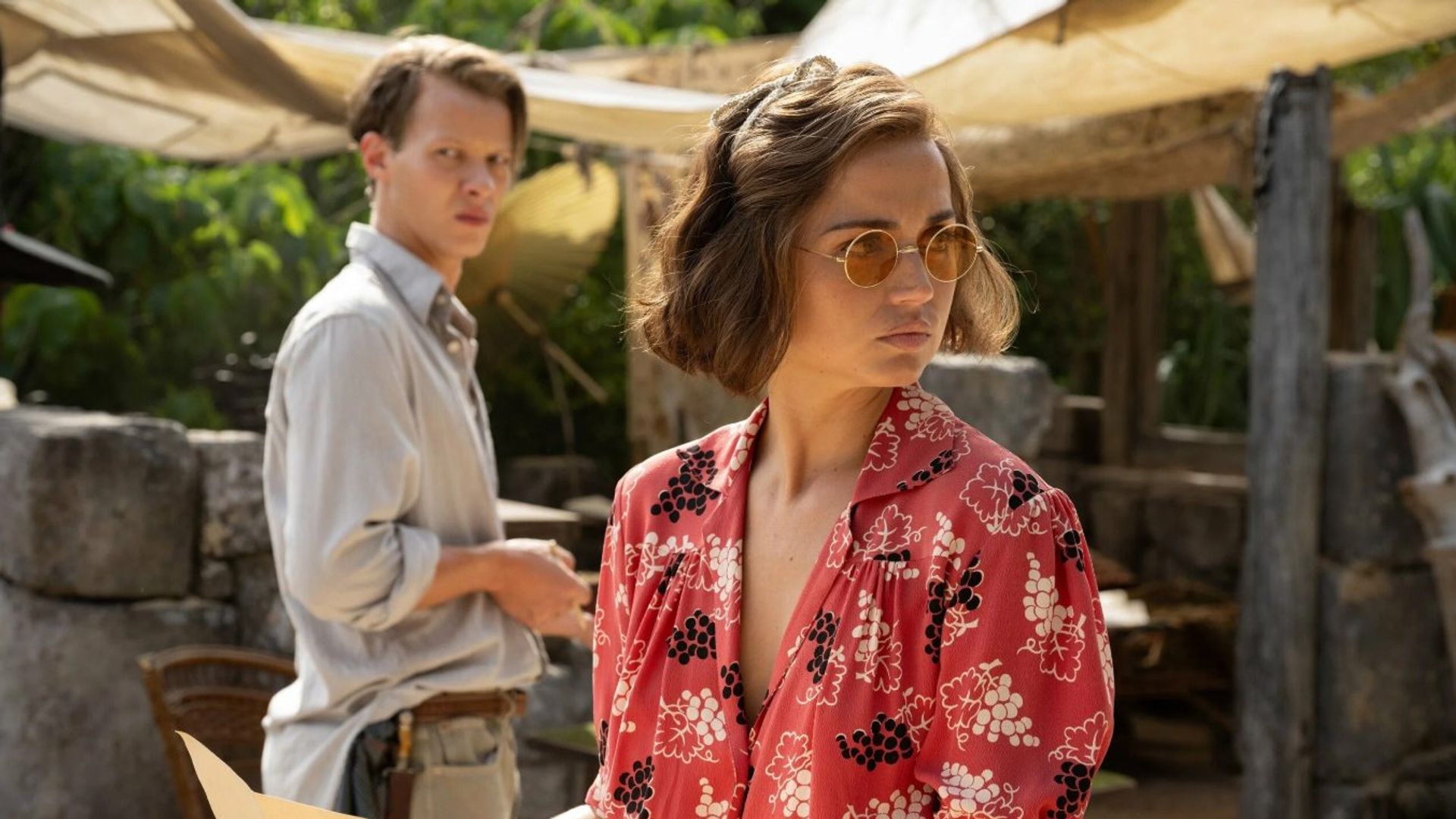 Ana de Armas con una blusa roja estampada y gafas de sol sostiene un mapa, parada junto a un hombre con una camisa clara, la escena ambientada contra ruinas arqueológicas
