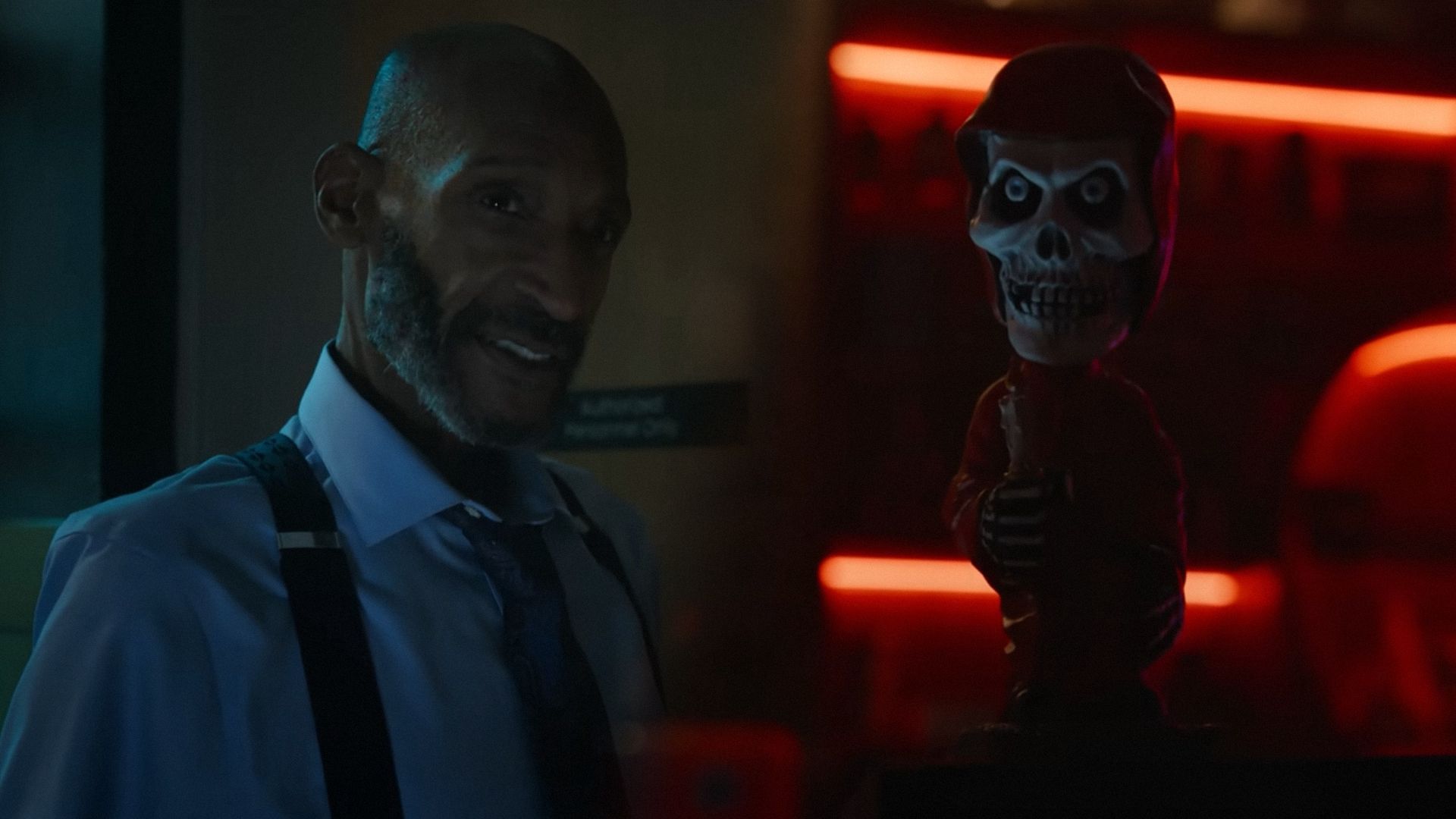 El actor Tony Todd con una camisa blanca y tirantes está de pie en un ángulo y mira de lado contra el fondo de una oficina