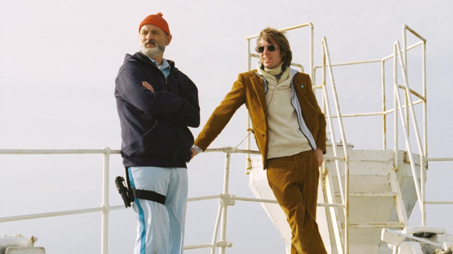 El actor Bill Murray con un sombrero rojo y el director Wes Anderson con una chaqueta marrón se sostienen en la barandilla del barco contra el cielo