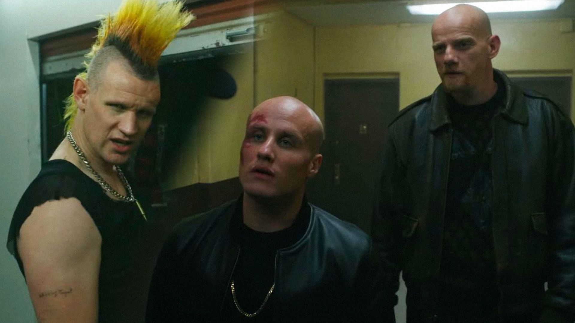 El actor Matt Smith con un mohawk mira hacia un lado, los actores Nikita Kukushkin y Yuri Kolokolnikov en trajes de cuero están en el pasillo y miran hacia un lado