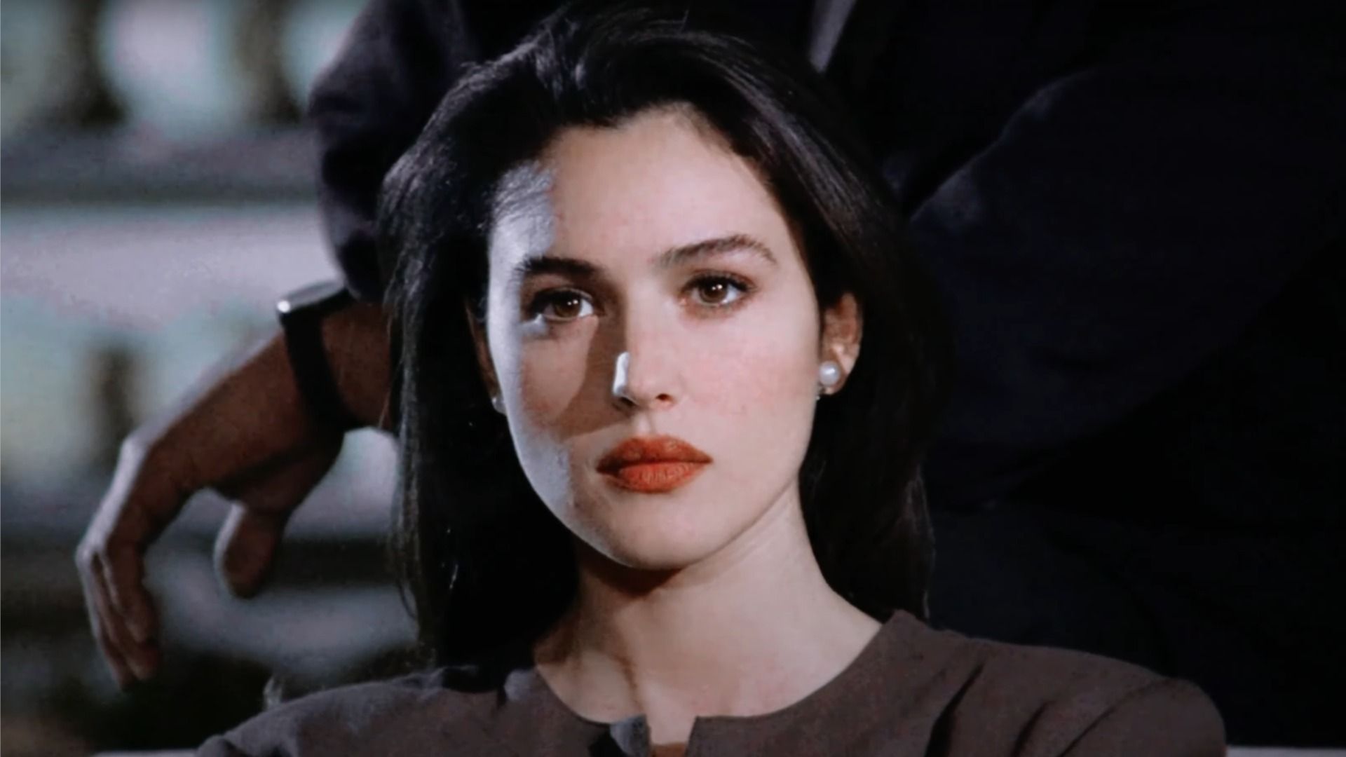 Actriz Monica Bellucci con lápiz labial rojo en los labios mira hacia un lado contra el fondo de un hombre en traje