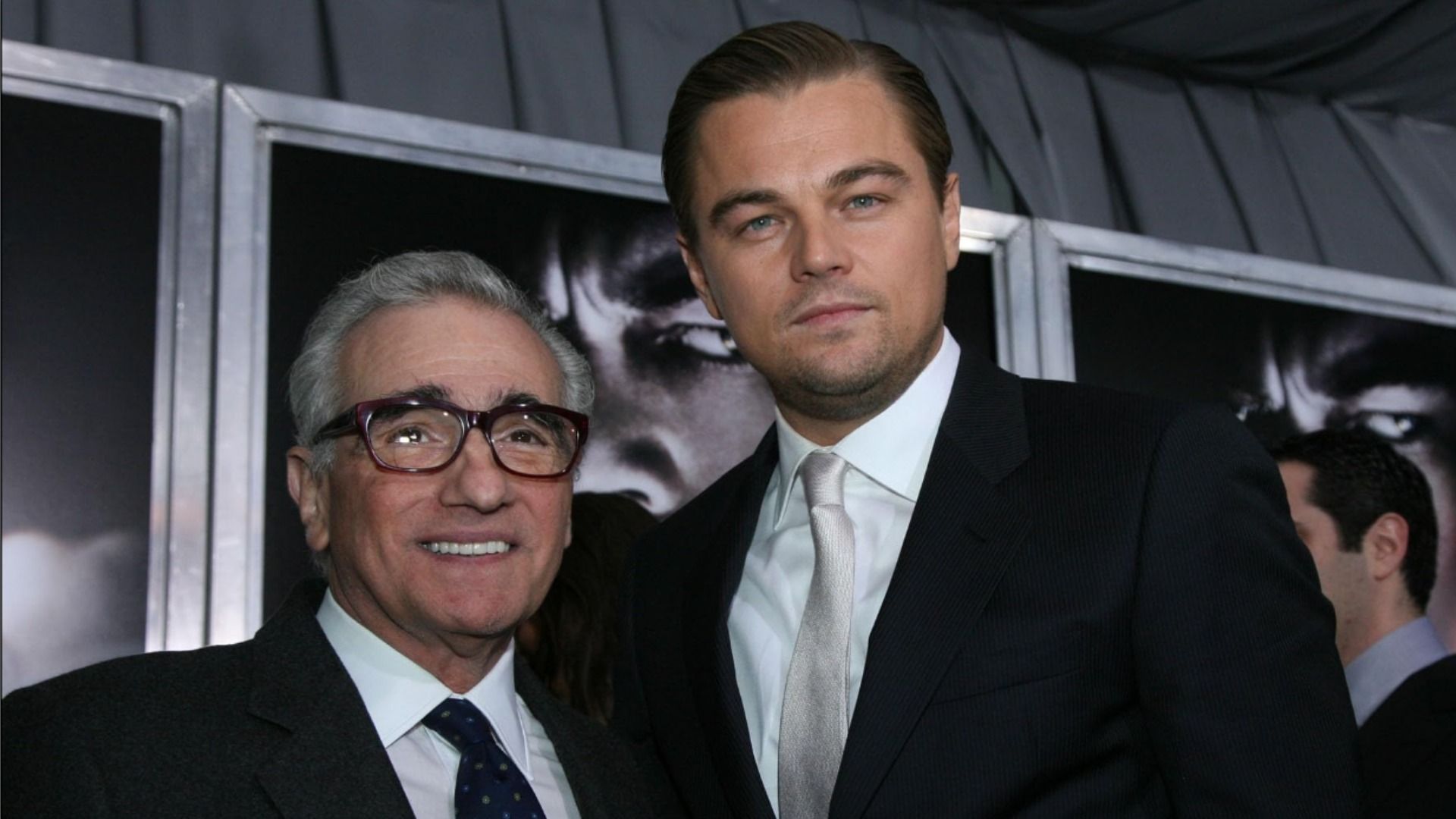 El director Martin Scorsese con gafas sonríe junto al actor Leonardo DiCaprio, ambos con trajes negros frente a un fondo de carteles