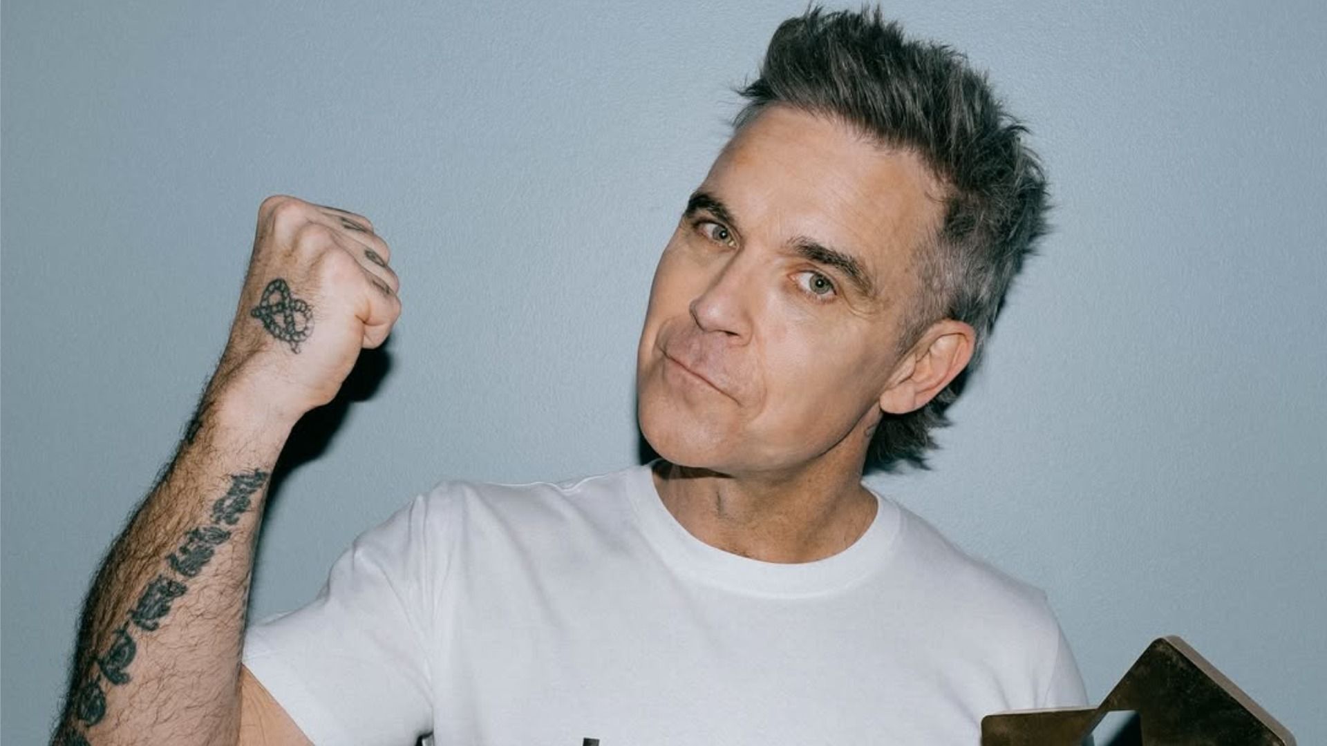 El cantante Robbie Williams levantó el puño y sonríe, mirando a la cámara contra un fondo gris