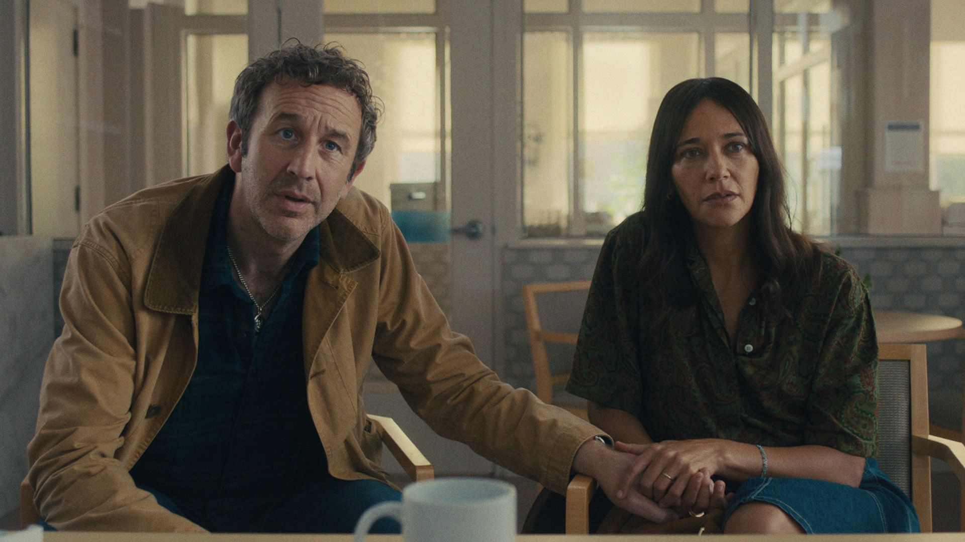 Chris O'Dowd y Rashida Jones sentados en sillas con expresiones preocupadas, tomados de la mano