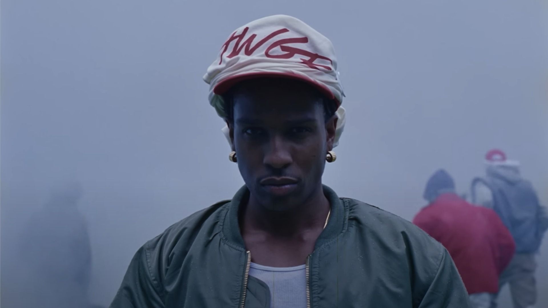 El rapero A$AP Rocky con una chaqueta bomber verde mira a la cámara contra el telón de fondo de una calle humeante