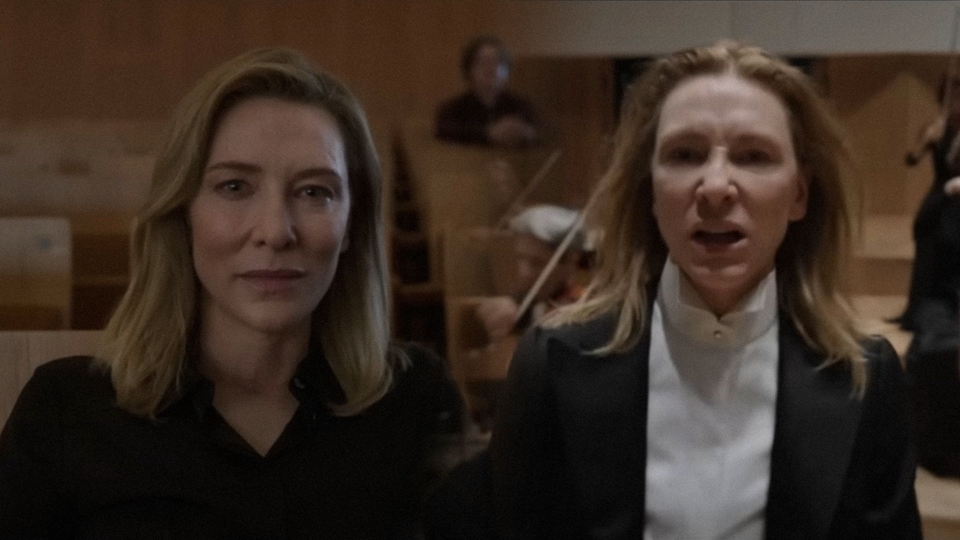 La actriz Cate Blanchett con traje negro mira amenazadoramente a la cámara con un telón de fondo de violinistas