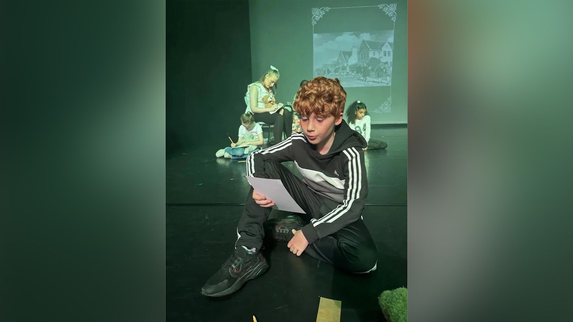 Alastair Stout con chándal sentado en un escenario teatral leyendo de una hoja