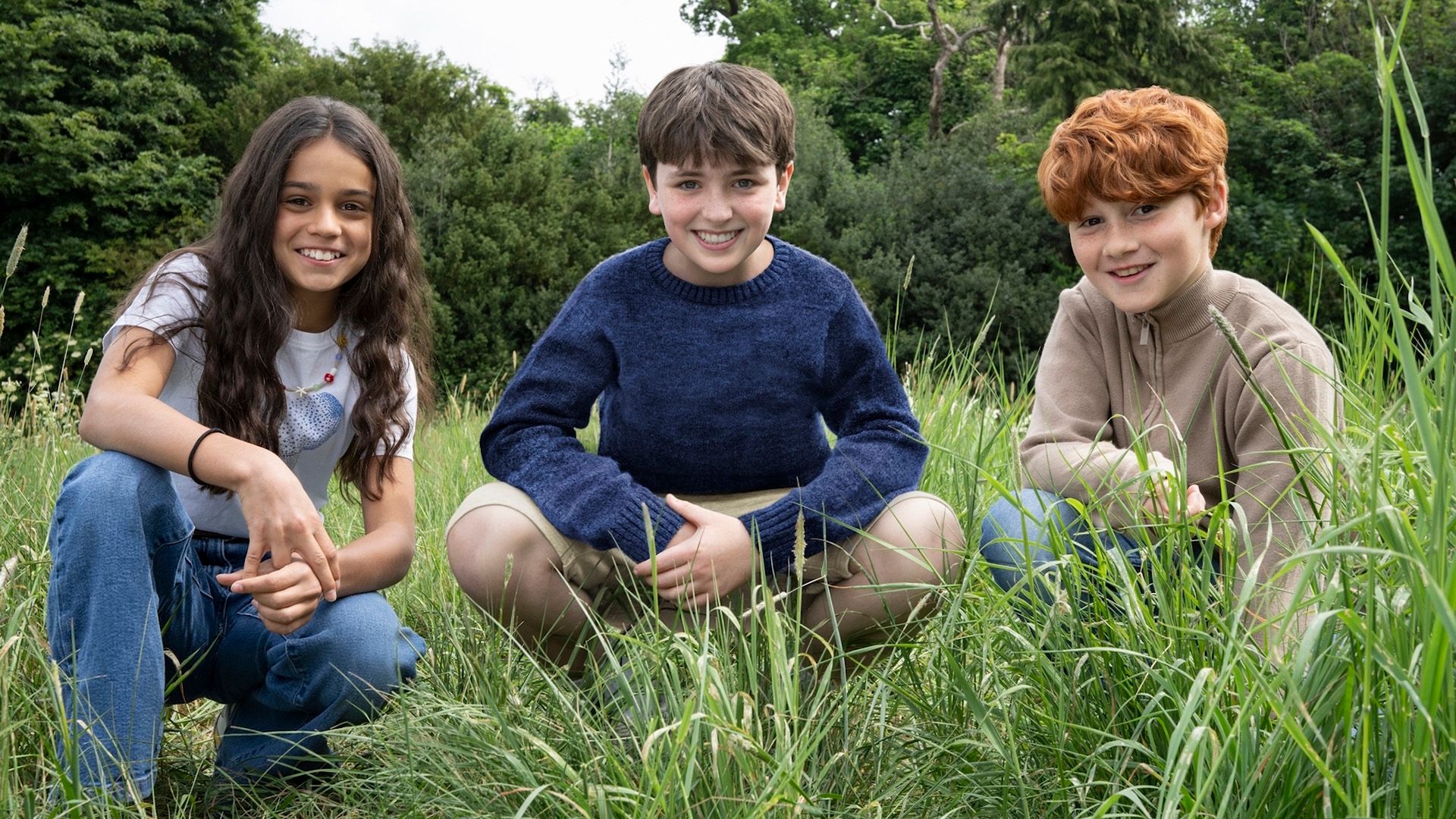 Los jóvenes actores Arabella Stanton, Dominic McLaughlin y Alastair Stout agachados en la hierba sonriendo
