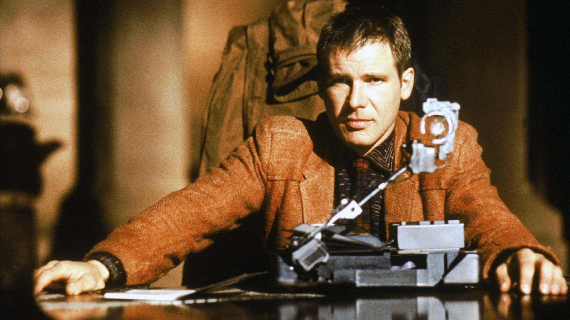 El actor Harrison Ford como Rick Deckard sentado en una mesa con chaqueta marrón