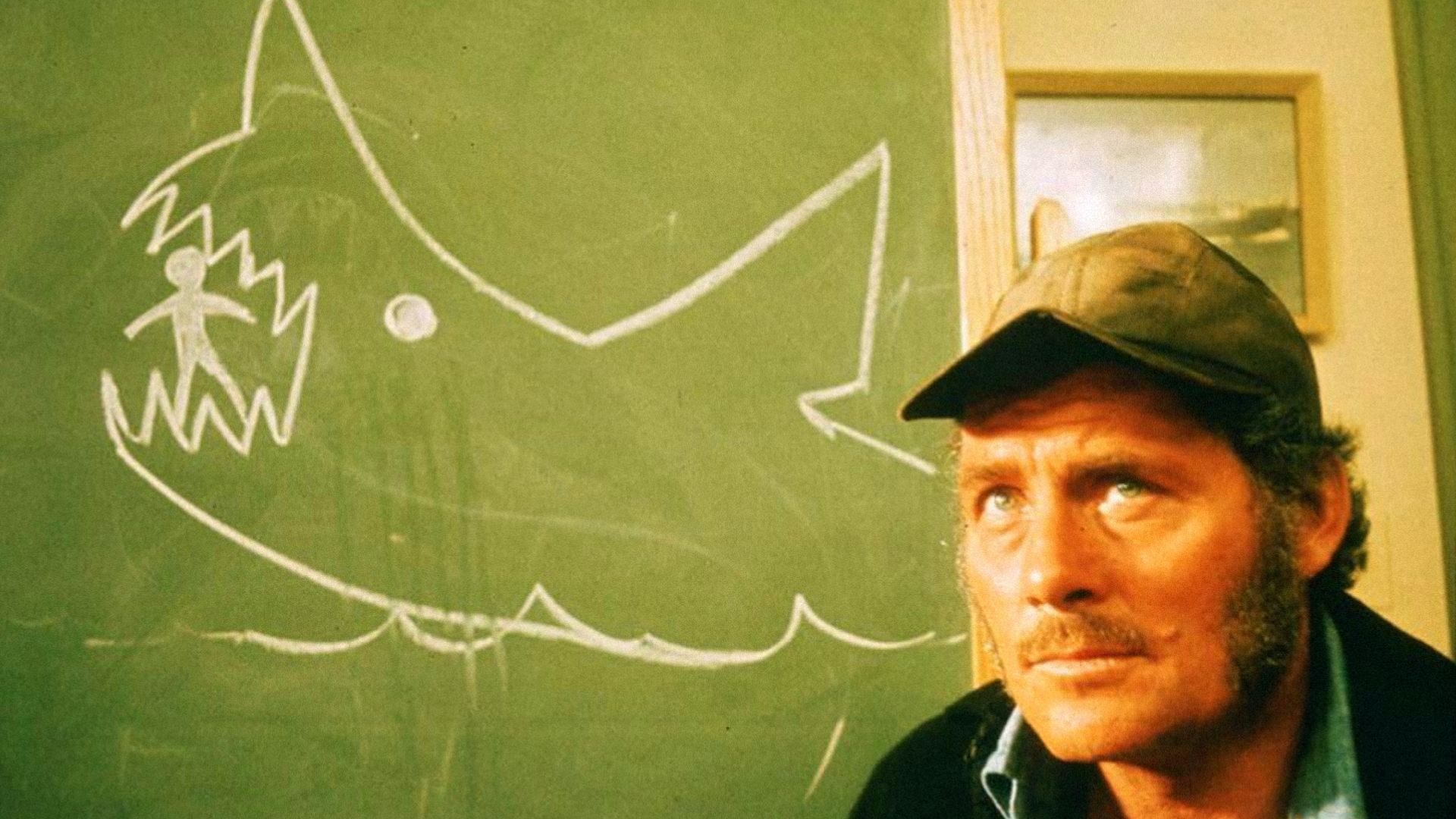 El actor Robert Shaw con una gorra gris de pie junto a una pizarra con un tiburón dibujado con tiza