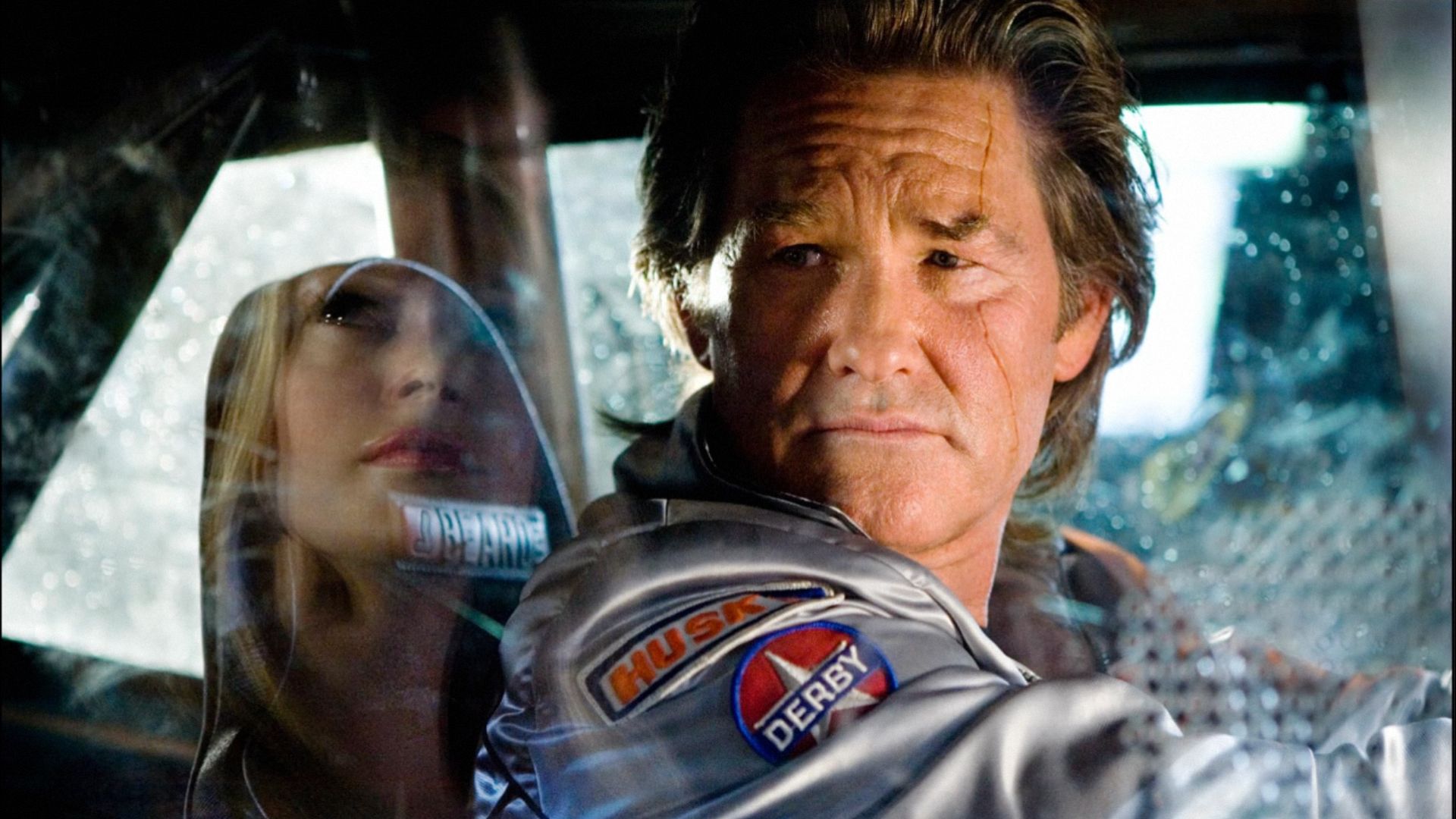 El actor Kurt Russell con chaqueta plateada sentado al volante de un coche mirando hacia un lado